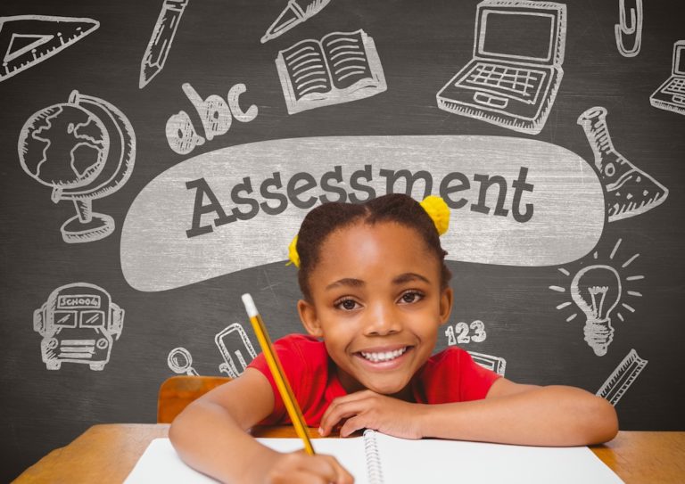 Young, smiling girl sitting at a desk with a pencil and paper with assessment written on the blackboard behind her.