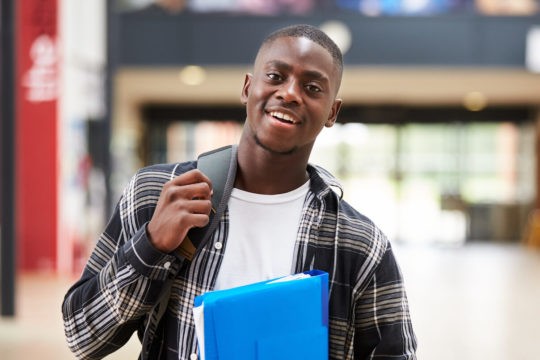 Young man standing with a backpack and a binder in a college hallway.