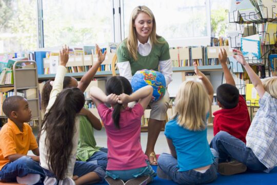 Students sit in front of their teacher who is talking; they are watching and listening intently.