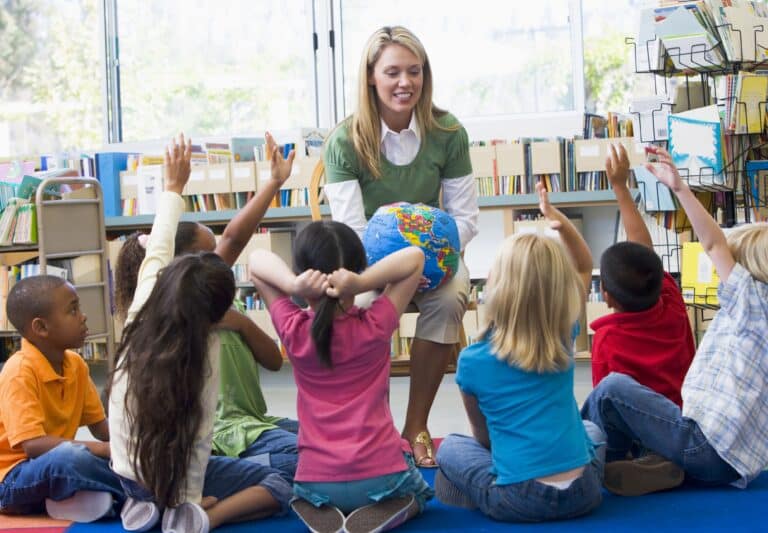 Students sit in front of their teacher who is talking; they are watching and listening intently.