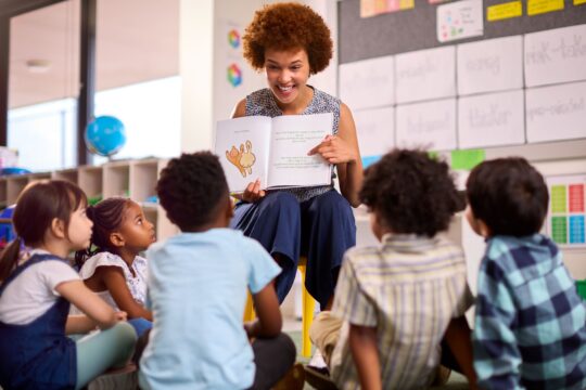 A teacher sits with her students while reading them and showing them a book.