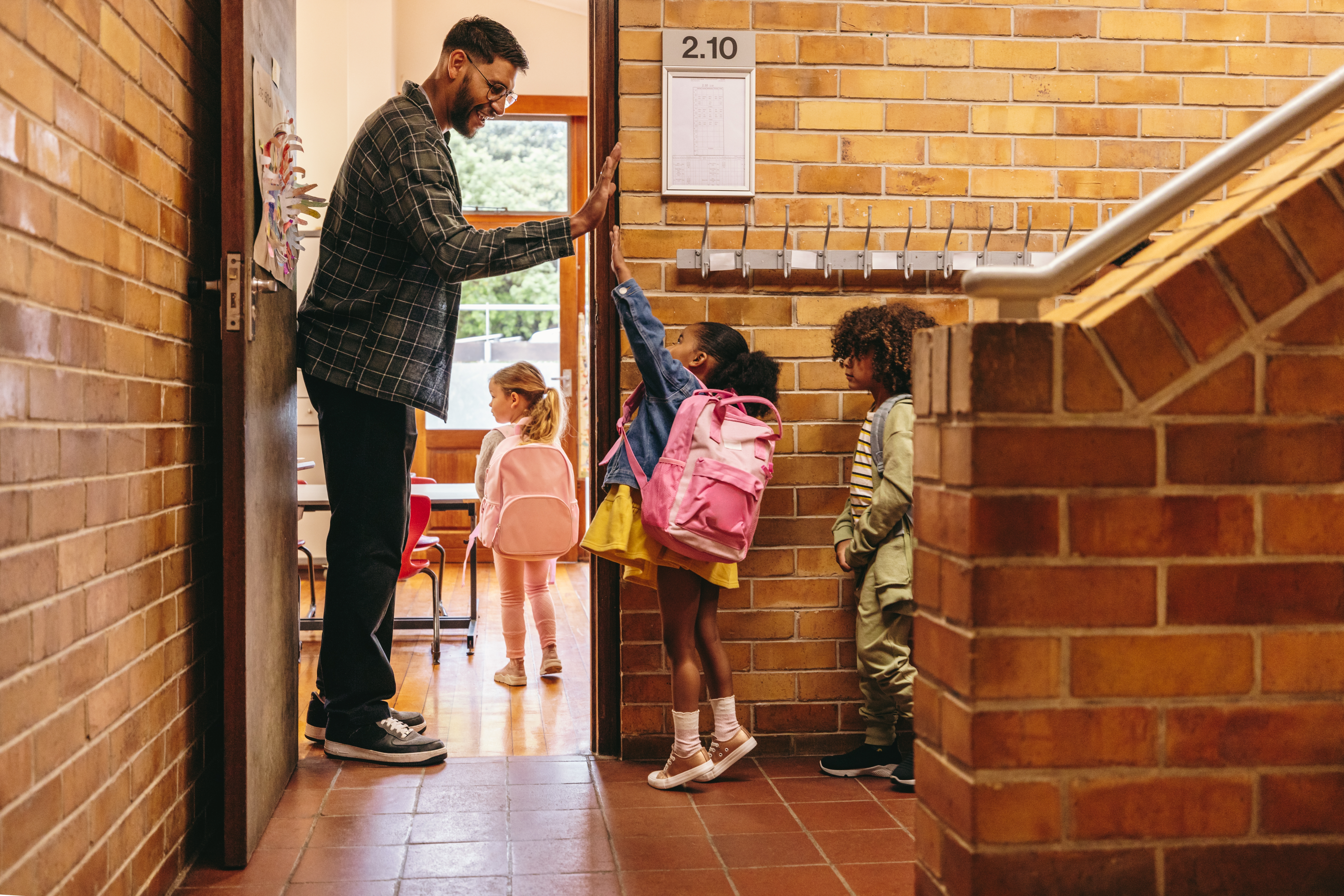 A teacher stands outside his classroom, waving to his students as they come inside.