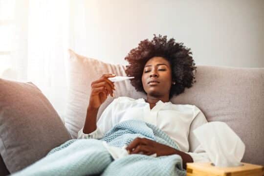 A woman lays in bed sick while looking at a thermometer, representing a sick day.