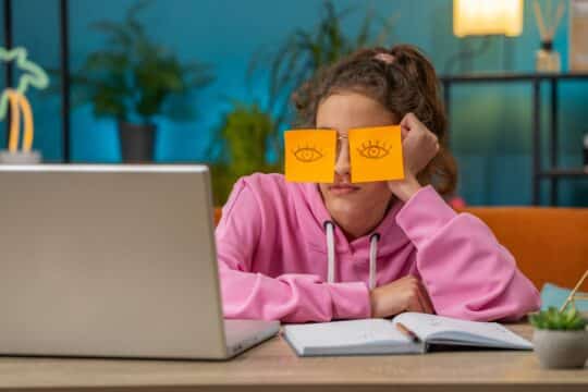 A student sits in front of her laptop, with post it notes over her eyes to make it look like she has her eyes open, representing student burnout.