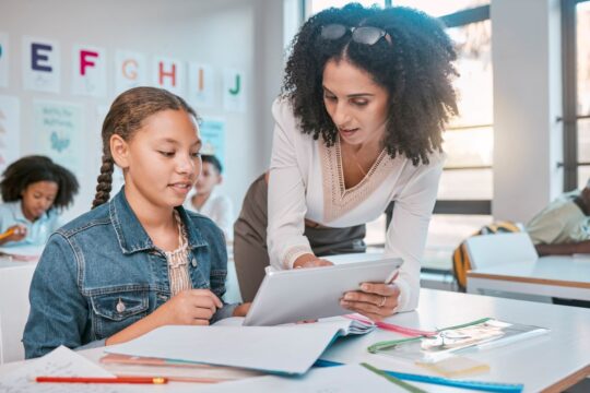 A teacher assisting a student with a digital tablet at a desk in a classroom. Focused on learning outcomes and assessment techniques.