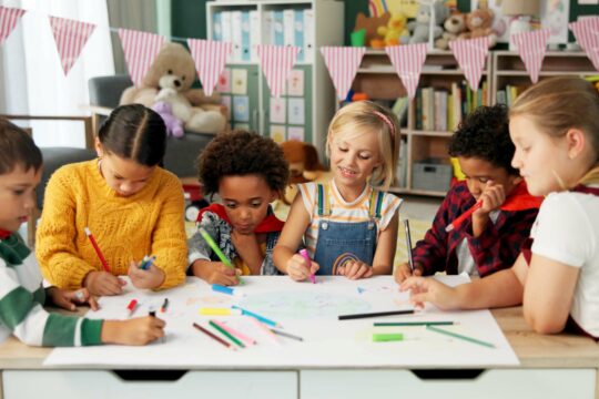 Art projects for students; young kids sit at a table, drawing with colored pencils on a big piece of paper.