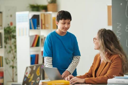 Restorative practies: a smiling student stands at his teacher’s desk, talking to her.