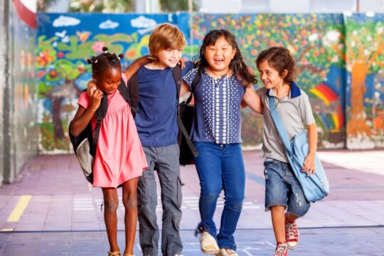Four multi-cultural children walk through school with their arms around each other.