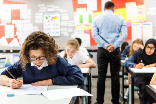 Students take a written exam in the classroom while their teacher walks around and checks on them.