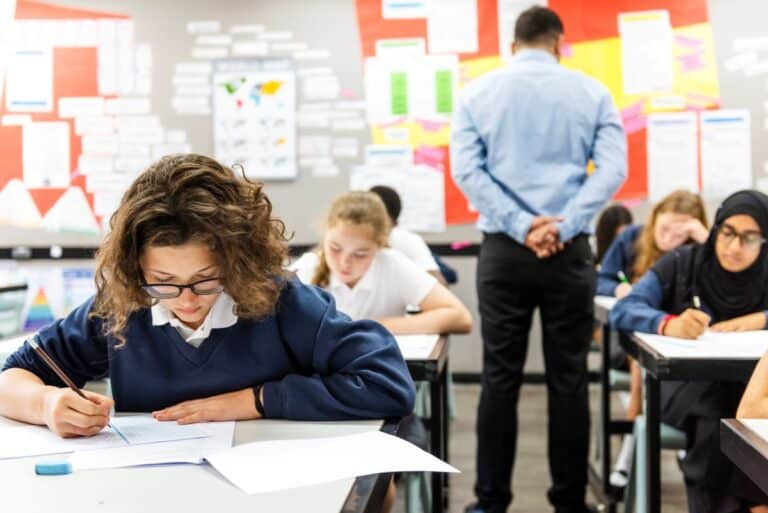 Students take a written exam in the classroom while their teacher walks around and checks on them.