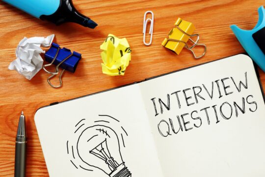 An open notebook has a lightbulb and “interview questions” written inside of it with crumpled pieces of paper around it.