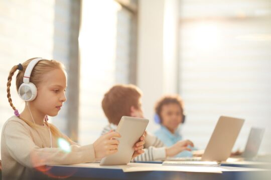 Three students sit at their school desks; one is on their tablet and the other two are on their laptops.